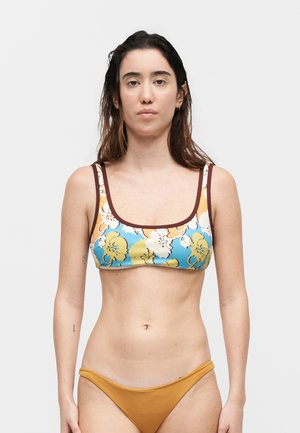 Γυναίκα με μακριά σκούρα μαλλιά που φοράει μπικίνι με λουλουδένιο σχέδιο επάνω μέρος και μονόχρωμο κάτω μέρος σε μουσταρδί, στέκεται μπροστά σε απλό φωτεινό φόντο.