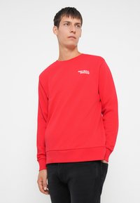 Piros pamut pulóver, crewneck stílusú, kis fehér "RED BULL RACING" logóval a mellkason, bordázott mandzsettákkal és aljjal.