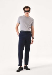 Moški, oblečen v sivo majico s kratkimi rokavi, temno modre hlače, črni pas, črni poslovni čevlji, črna sončna očala in ročna ura, stoji pred enostavnim ozadjem.