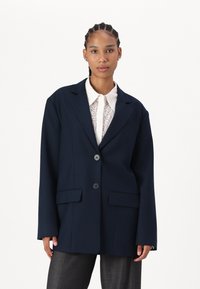 Marineblå oversized blazer laget av strukturert stoff, med to knapper, to frontlommer og krage med hakk, båret over en lys mønstret skjorte.