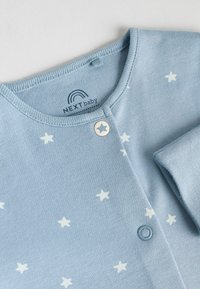 Lyseblå babycardigan lavet af blødt stof, med hvide stjernemønstre, en rund halsudskæring og trykknaplukning.