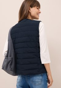 Gilet imbottito blu navy con design trapuntato, tasca con zip a sinistra, indossato sopra una camicia a maniche lunghe bianca, abbinato a jeans di denim.
