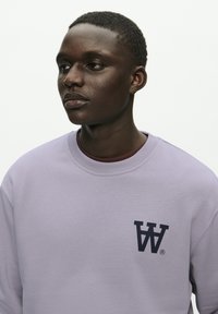 Light purple sweatshirt made from soft fabric, with a crew neckline and a dark blue logo featuring stylized letters on the chest.