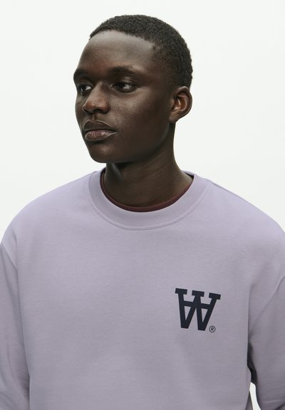 Sweat-shirt lilas clair en tissu doux, avec un col rond et un logo bleu foncé comportant des lettres stylisées sur la poitrine.
