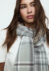 Écharpe à carreaux grise en tissu doux, avec un motif à damier aux accents blancs et bruns, drapée de manière décontractée autour du cou.