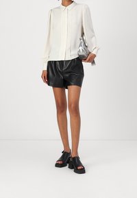 Weißes, plissiertes Blusentop mit Kragen, kombiniert mit schwarzen Leder-Shorts. Schwarze Plateausandalen und eine metallische Handtasche runden den Look ab.