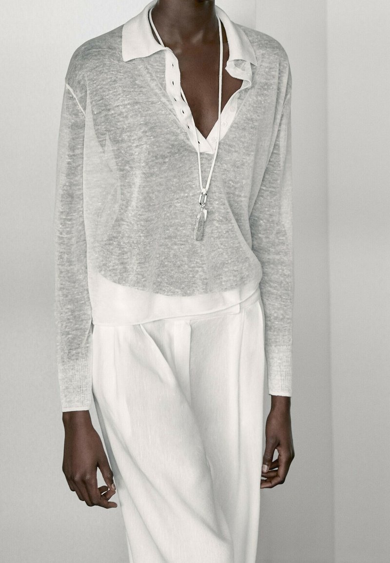 Personne portant un haut gris transparent à manches longues par-dessus une chemise blanche à col et un pantalon blanc ample, avec un long collier pendentif.