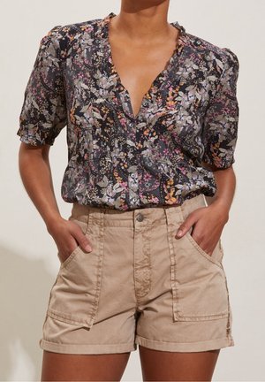 Kvinde iført en mørk blomstret knapp-up bluse med korte ærmer, stoppet ned i beige højtaljede shorts med store forlommer.