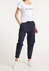 Camiseta blanca de manga corta con estampado "DreiMaster", combinada con pantalones cargo azul marino que presentan bolsillos laterales y puños elásticos, y llevada con zapatillas blancas.