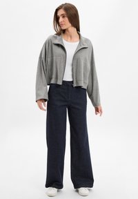 Graue cropped Jacke mit hohem Kragen, Frontreißverschluss, kombiniert mit lockeren, dunkelblauen Jeans und weißen Sneakers. Der Stoff hat eine glatte Textur.