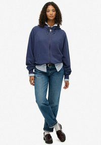 Sudadera con cremallera en navy y capucha con cordón, llevada sobre una camisa a rayas, combinada con jeans azules holgados y zapatos marrones. El modelo se encuentra de pie.