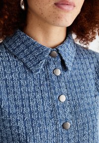 Chemise bleu texturée à manches longues avec un col, présentant des coutures visibles et des boutons métalliques sur le devant.
