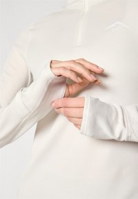 Svijetlosmeđa košulja dugih rukava s rebrastom teksturom, polu-zip ovratnikom i otvorima za palčeve; sadrži mali logotip na prsima.