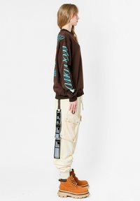 Sudadera marrón con letras turquesa, pantalones cargo beiges con bolsillos laterales y correas, y botas naranjas con una suela gruesa y texturizada.