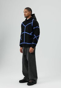 Zwarte hoodie met een blauw geometrisch patroon, voorzien van een grote voorkantzak en een capuchon met trekkoord, gecombineerd met grijze wijde jeans en zwarte schoenen.