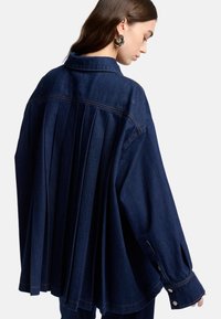 Mørkeblå denimjakke med plisseret rygdesign, lange ærmer og knaplukning ved manchetterne. Synlige syninger og krave.