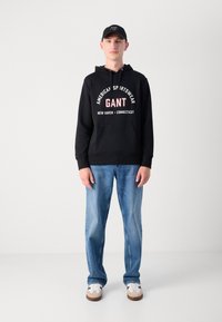 Čierna mikina s kapucňou s logom "GANT" v červenej a bielej farbe, kombinovaná s svetlomodrými džínami a bielymi teniskami, doplnená o zladenú čiernu čiapku.
