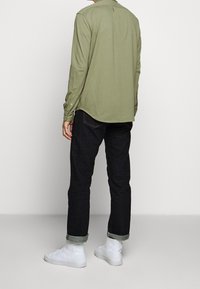 Muž stojící zády, oblečený v olivově zelené košili s dlouhým rukávem, tmavých džínech se zakouřenými nohavicemi a bílých vysokých teniskách na jednofonním pozadí.