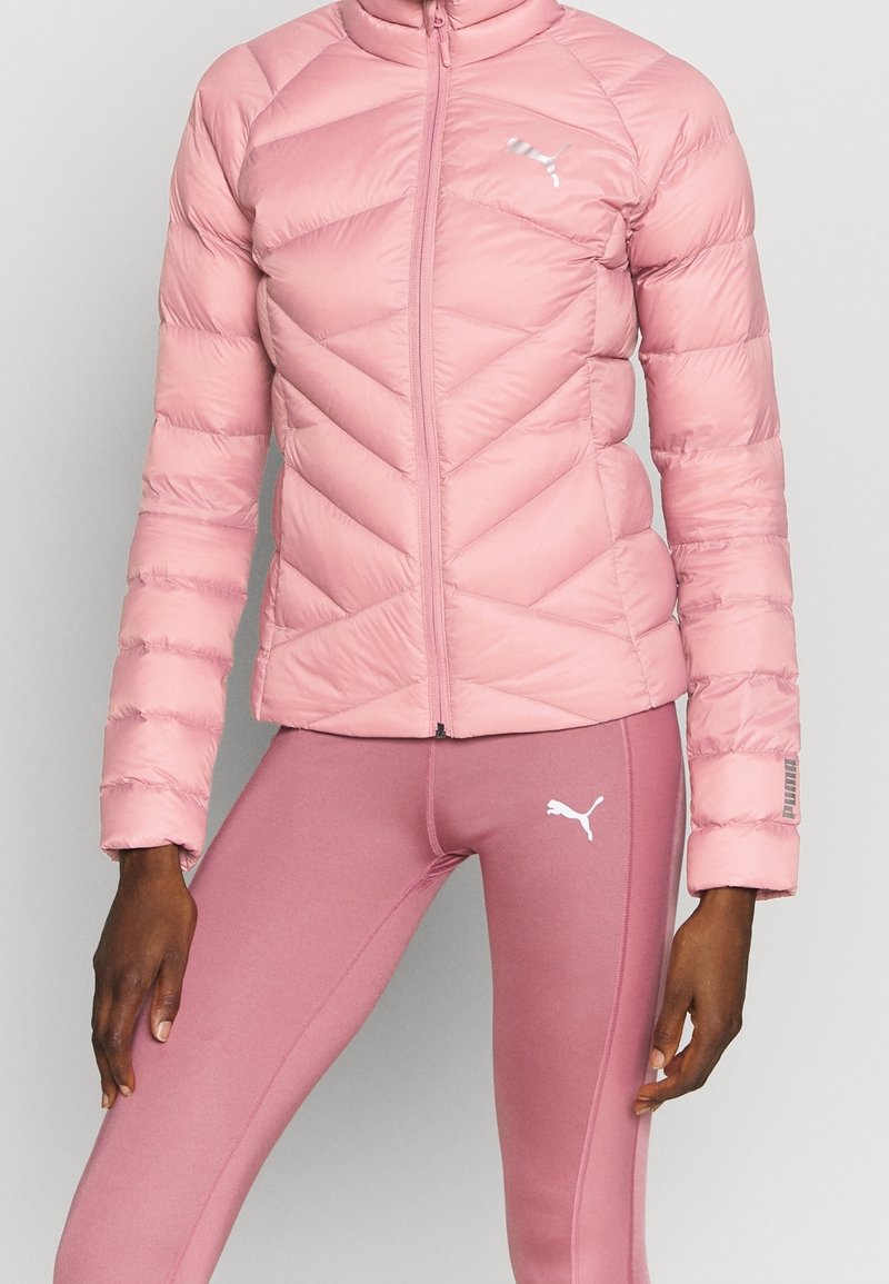 Femme portant une veste matelassée rose et des leggings roses assortis, tous deux arborant le logo Puma, debout avec les bras détendus le long du corps.