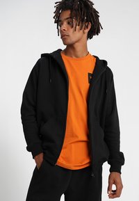 Sudadera negra con cremallera sobre una camiseta naranja, con un bolsillo frontal, capucha con cordones y un pequeño logo en el pecho de la camiseta.