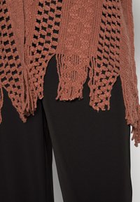 Szydełkowy szal w kolorze przybrudzonego różu z otwartymi wzorami, z frędzlami na brzegach; nałożony na czarny materiał, ukazujący fakturę i skomplikowane przeszycia.