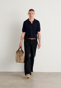 Marine polo shirt og sorte bukser båret av en person som holder en tan duffel bag med brune detaljer. Glatt stoff, avslappet passform, minimalistisk design.
