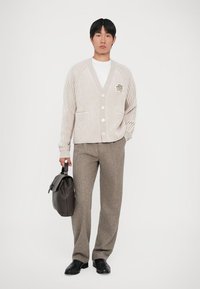 Beige geribde cardigan met v-hals, knoopsluiting en logo-detail, gecombineerd met bruine textuurbroek en een zwarte leren tas.