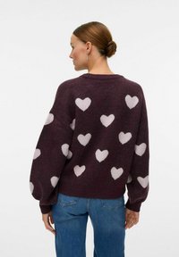 Maglione amaranto con motivi a cuore bianchi, morbido al tatto, vestibilità ampia, scollatura rotonda e polsini a coste. Abbinato a jeans blu.