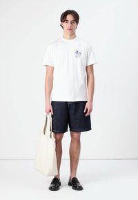 Hvit bomulls t-skjorte med en liten blå grafikk på venstre bryst, paret med mørke denimshorts. Modellen holder en beige totebag.