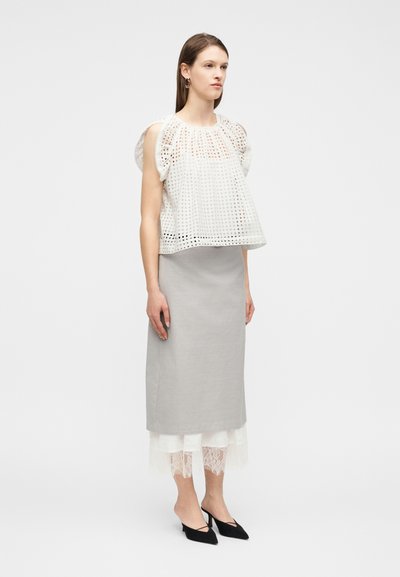 Femme portant un haut sans manches en dentelle ajourée blanche, une jupe midi gris clair avec un ourlet en dentelle blanche, et des escarpins noirs à bride arrière pointus, debout sur un fond blanc.