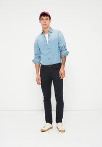 Lys blå denimskjorte, mørke slim-fit bukser og hvite joggesko med beige såler. Skjorten har knappestolpe foran og oppbrettede ermer.