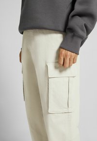 Beige cargobyxor i bomull med flera sidofickor, med rak skärning och en grå sweatshirt synlig vid midjan.