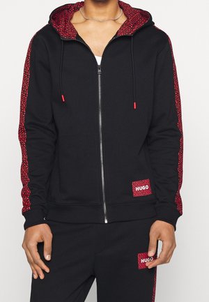 Zwarte zip-up hoodie met een rode patroonvoering en accenten. Beschikt over rode trekkoorden en zijstrepen. "HUGO" logo op de zak.