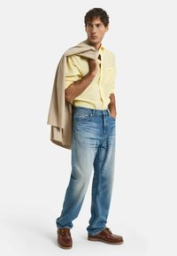 Informele outfit bestaande uit een gele overhemd, lichtblauwe jeans en bruine bootschoenen. Een beige trui is over één schouder gedrapeerd.