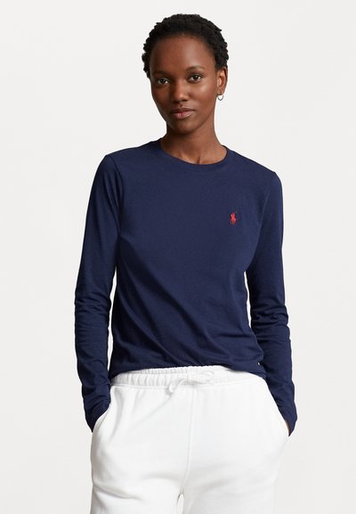 Chemise à manches longues bleu marine en coton avec un col rond et un petit logo rouge sur la poitrine. Associée à un pantalon de survêtement blanc.