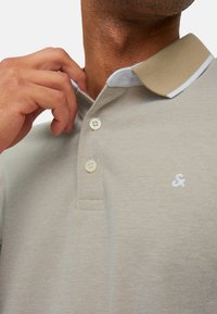 Harmaa polo-paita, jossa on vaaleanruskea kaulus, kolme nappia ja pieni valkoinen logo rinnassa. Paidassa on teksturoidun kankaan kuvio.