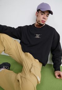 Mladý muž s vitiligo, který má na sobě fialovou čepici, černou mikinu Timberland, béžové cargo kalhoty a černé tenisky, sedí na zelené podlaze.
