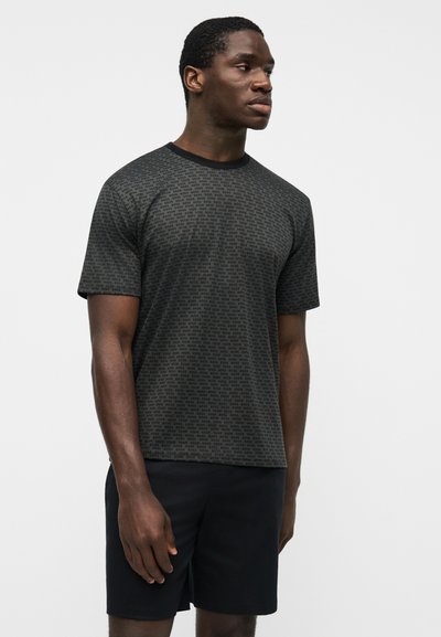 Homme portant un T-shirt gris foncé à motifs et un short noir, debout et regardant vers la droite sur un fond clair uni.