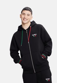 Felpa nera con zip realizzata in un misto cotone, dotata di un cappuccio con lacci verde e rosso, polsini a coste con strisce coordinate e dettaglio del logo.