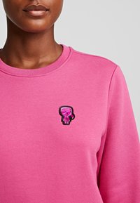 Sudadera rosa con un pequeño y brillante figura de dibujos animados en rosa y negro, con gafas de sol bordada en el lado izquierdo del pecho.