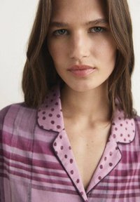 Top de cuadros púrpura y rosa con un cuello de puntos, tejido ligeramente texturizado y escote abotonado. El diseño incluye rayas y acentos en contraste.