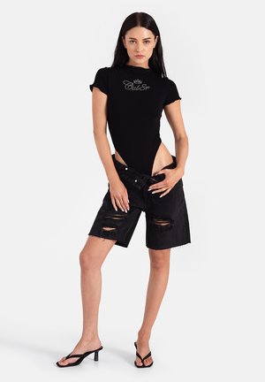 Czarny body z krótkimi rękawami i logo z kryształków, w zestawieniu z czarnymi, postrzępionymi szortami dżinsowymi. Modelka ma na sobie czarne klapki.