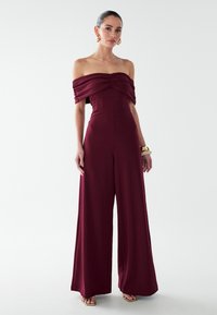 Moteris vilki bordo spalvos off-shoulder kombinezoną su plačiomis kojomis, auksinėmis auskaromis žiedais ir apyrankėmis, stovinti priešai paprastą baltą foną.