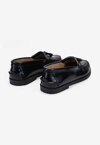 Mocasines de charol negro con un acabado suave, que presentan acentos de borlas, costuras decorativas y una suela baja con textura.