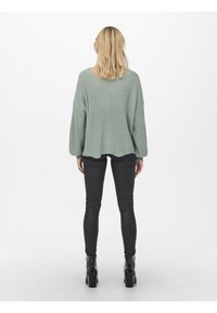 Pull en maille oversize vert clair avec épaules tombantes, associé à un pantalon skinny noir et des bottines noires. Texture lisse, coupe décontractée.