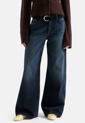 Persona che indossa jeans scuri a gamba larga con cintura nera, maglione marrone a maniche lunghe e scarpe basse giallo chiaro.