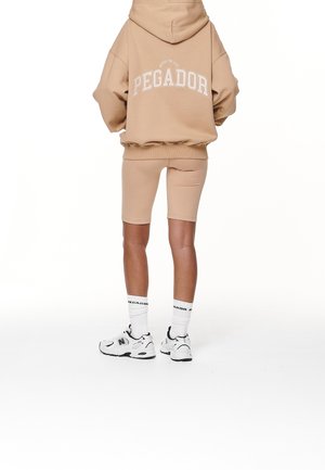 Person wearing beige oversized hoodie with "PEGADOR" text, matching bike shorts, white socks, and white sneakers, viewed from the back.