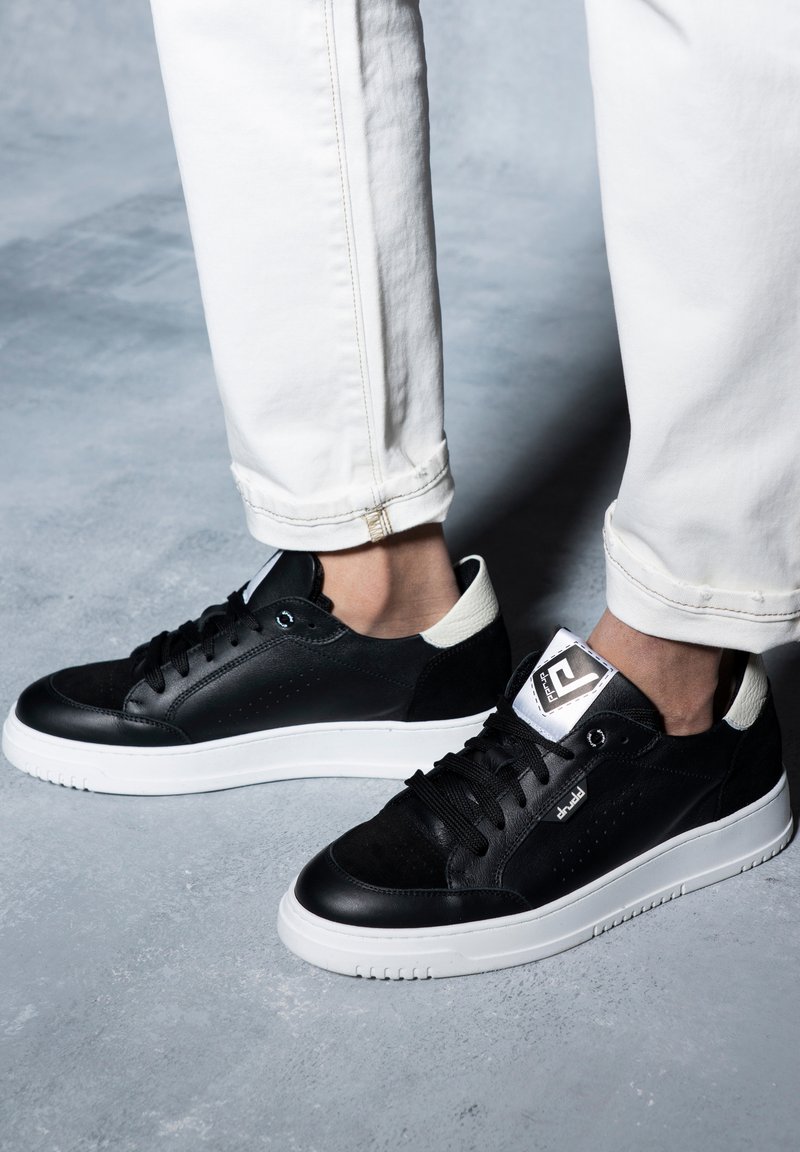 Sneaker in pelle nera con finitura liscia, suola in gomma bianca e dettagli del tallone in contrasto. Presenta lacci tondi e un design superiore perforato.