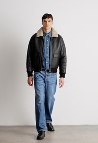 Black leather bomber jacket with a tan shearling collar, paired with a denim button-up and light blue denim jeans. Black shoes.