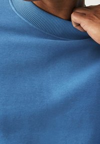 Felpa blu con collo a girocollo a coste, texture liscia e cuciture visibili. Messa a fuoco ravvicinata sui dettagli del tessuto e del colletto.